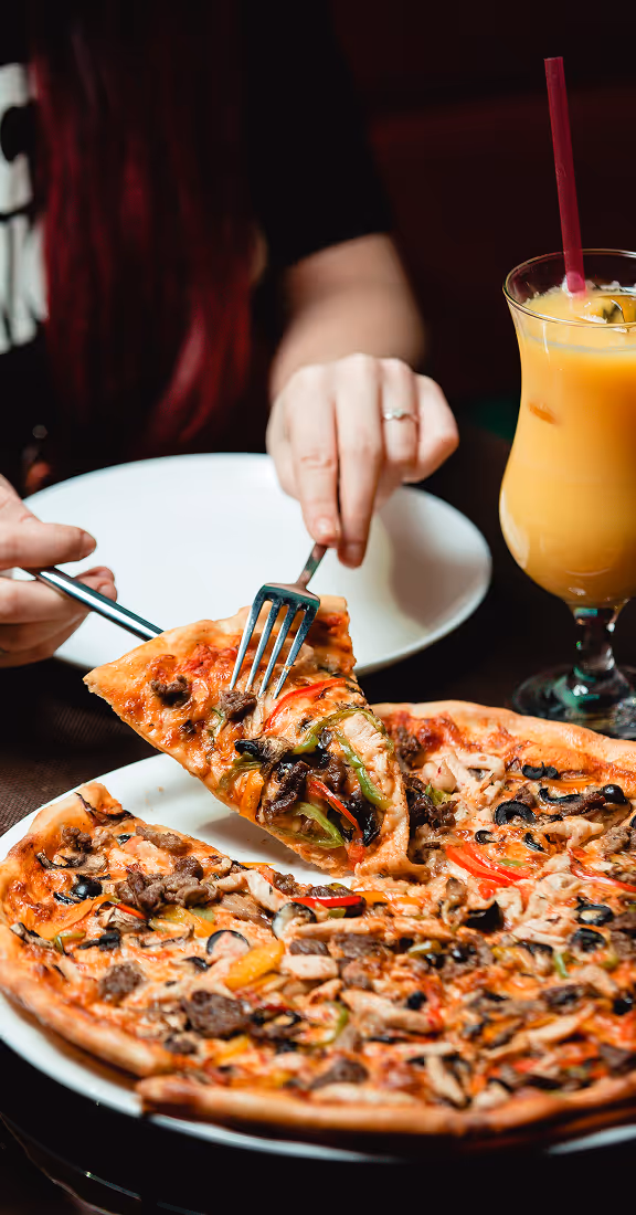 Person holding a fork and knife cutting a slice of vegetable and meat pizza with a glass of orange juice with a straw beside the plate.